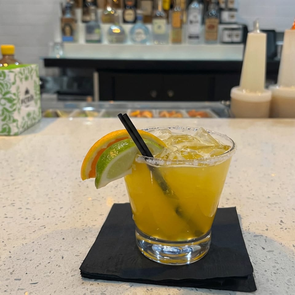Golden citrus cocktail with ice, black straw, and orange and lime wedges on a black napkin at a restaurant bar.