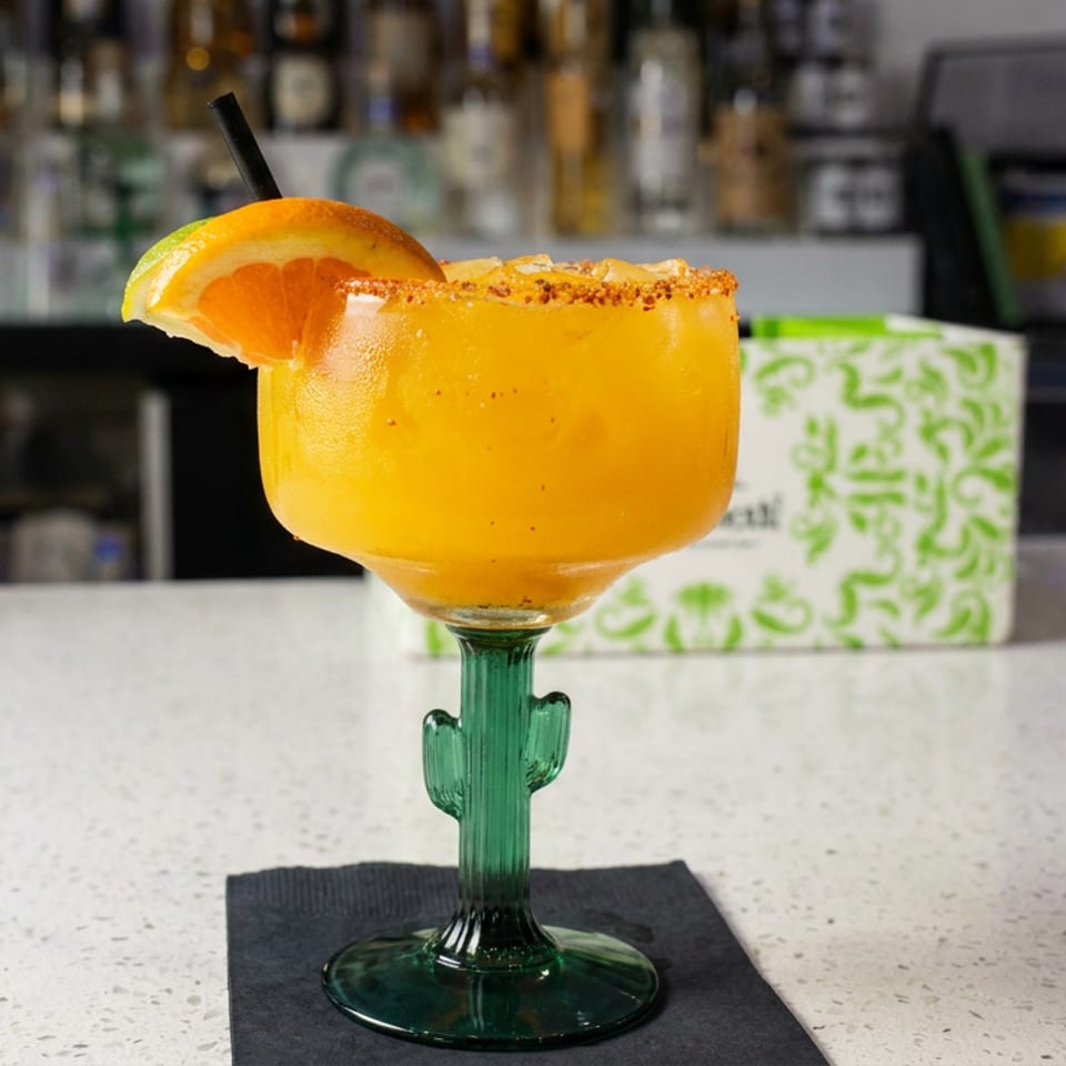 Bright orange cocktail with a salted rim and orange garnish served in a green cactus-shaped glass at a bar.