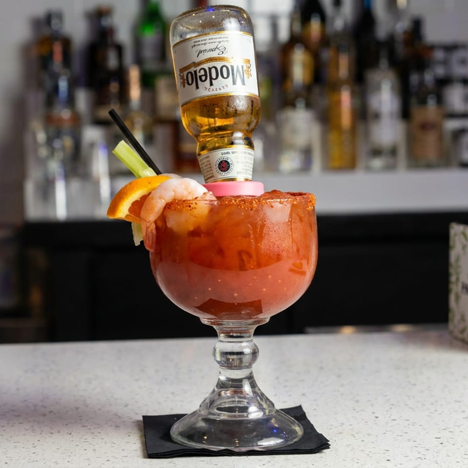 Spicy shrimp cocktail garnished with citrus and celery, served in a glass with an upside-down Modelo beer bottle at a bar.