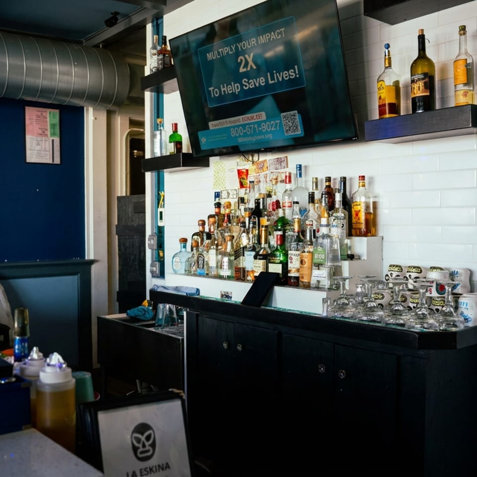 Restaurant bar with liquor bottles, glassware, beer taps, and a television displaying a charity message.