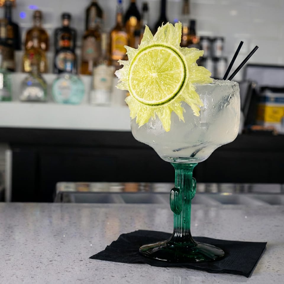 Frosted cocktail in a green-stemmed glass, garnished with a lime wheel, sits on a black napkin at a bar.